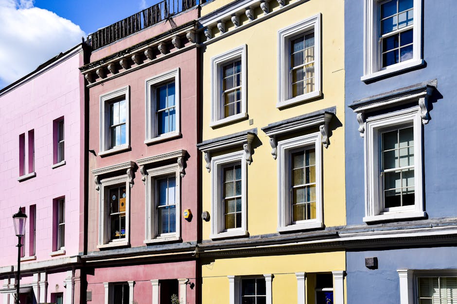 A row of three colourful residential buildings with distinct facades, featuring white-framed sash windows and decorative cornices. The building on the left is painted in pastel pink, while the central building in the middle is painted pale yellow, and the one on the right is a soft blue. Each set of windows has detailed molding beneath them, and some windows have partially closed blinds or curtains visible inside. The buildings are set against a partly cloudy sky, with a traditional black street lamp mounted on a pole visible at the left edge of the image. The scene suggests an urban streetscape, possibly in a densely built residential area. The clean and well-maintained appearance of the facades indicates a tidy environment, which aligns with efforts of professional waste clearance services such as Waste Clearance Notting Hill, to maintain cleanliness and proper waste management in the area. No rubbish or waste materials are visible in the image, emphasizing an emphasis on property maintenance and environmental care typical of private rubbish collection and on-site clearance operations.