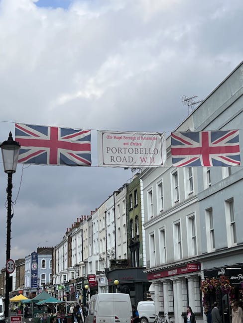 A street scene in Notting Hill featuring a row of closely packed Victorian-style terraced buildings with facades painted in white, pale green, and other soft tones. The sky above is overcast with grey clouds. In the foreground, there is a black street lamp and a hanging banner displaying the Union Jack flags on either side, with a central sign reading 'Portobello Road W11' and indicating the area is part of the Royal Borough of Kensington and Chelsea. Below the banner, the street is active with pedestrians, small open-air market stalls with colorful umbrellas, and parked white vans. The scene captures a lively, urban environment, typical of the area known for its markets and independent shops, representing an example of private waste management and rubbish removal activities that support local commerce and community events. Waste Clearance Notting Hill specializes in rubbish removal, often assisting with clearance of commercial and residential waste from this vibrant street.