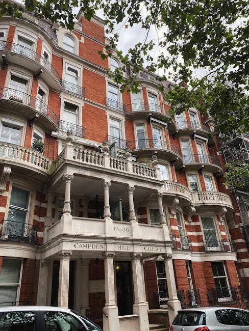 A multi-story red brick residential building with ornate white stone decorative elements and curved balconies, located on Campden Hill Court. The building's facade features large windows with white frames, some partially obscured by tree branches with green leaves in the foreground. The entrance is marked by a portico with four white columns supporting a balcony above, and the address number '13-36' is visible on the entrance structure. Parked cars are seen at the street level in front of the building. The scene appears to be during daylight with natural light illuminating the structure, and the overall presentation suggests a well-maintained property within an urban environment. The image subtly relates to private waste handling by showing a residential property that may require rubbish removal or clearance services, typical of independent collection options in the area, as provided by Waste Clearance Notting Hill.