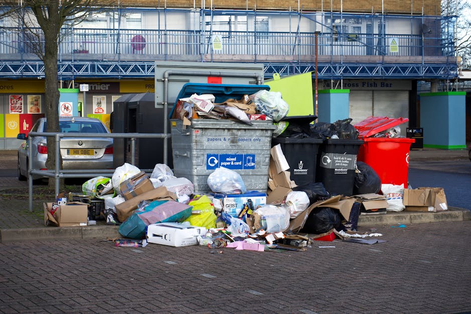 A large collection of mixed waste, including cardboard boxes, plastic bags, paper, and packaging materials, is piled outside a commercial building near a parking area. The rubbish is scattered across the sidewalk and street, with some items leaning against a metal railing directly in front of a silver hatchback car with a visible registration plate. Among the waste, there are black and green waste bins, with the open bin lid revealing more discarded paper and packaging. Behind the rubbish, a building with a blue metal scaffold and safety signage is visible, indicating ongoing construction or maintenance work. The scene is outdoors in a busy urban area, illuminated by natural daylight, with minimal greenery present. Waste Clearance Notting Hill often handles such waste, representing an example of private rubbish removal services aligned with alternative waste handling options like on-site clearance or independent collection outside standard municipal collections.