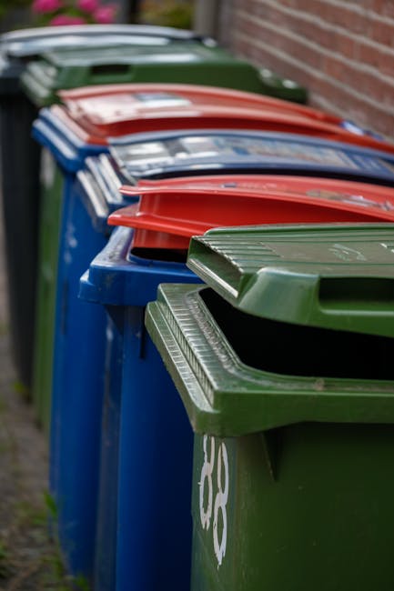A large collection of mixed waste, including cardboard boxes, plastic bags, paper, and packaging materials, is piled outside a commercial building near a parking area. The rubbish is scattered across the sidewalk and street, with some items leaning against a metal railing directly in front of a silver hatchback car with a visible registration plate. Among the waste, there are black and green waste bins, with the open bin lid revealing more discarded paper and packaging. Behind the rubbish, a building with a blue metal scaffold and safety signage is visible, indicating ongoing construction or maintenance work. The scene is outdoors in a busy urban area, illuminated by natural daylight, with minimal greenery present. Waste Clearance Notting Hill often handles such waste, representing an example of private rubbish removal services aligned with alternative waste handling options like on-site clearance or independent collection outside standard municipal collections.