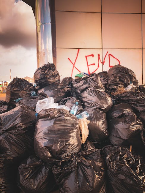 A large pile of black rubbish bags filled with waste is stacked against a modern tiled wall on an outdoor pavement area. The bags appear to contain mixed household refuse, with some slightly torn open, revealing plastic bottles and smaller debris inside. Several discarded plastic water bottles are visible, protruding from the bags or resting on top. In the background, graffiti with red spray paint spelling 'XEND' is visible on the wall, and a cloudy sky with hints of sunlight is seen above the scene. The setting suggests an instance of unauthorised dumping or a completed private rubbish clearance, with Waste Clearance Notting Hill potentially involved in the removal of this waste. The overall environment indicates an urban area requiring specialist waste removal services, with the scene highlighting the importance of professional rubbish collection and disposal for such waste accumulation.