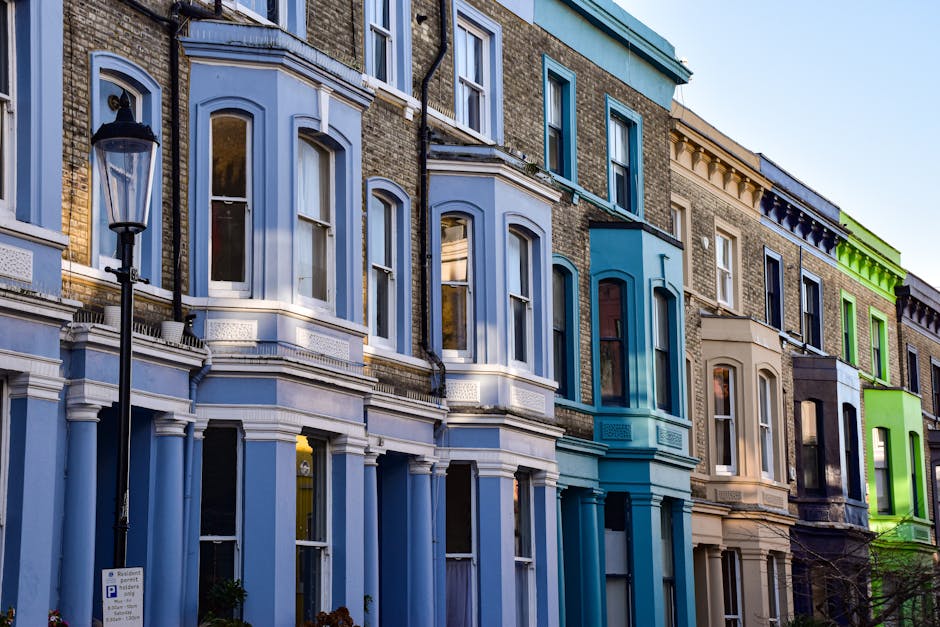 The image shows a row of Victorian-style terraced houses with bay windows, brick facades, and decorative architectural details. The buildings are painted in pastel and vibrant shades, including pale blue, light grey, beige, dark blue, and bright green, creating a colorful streetscape. Each house has large, multi-pane sash windows with white frames, some of which are partly open, revealing interior curtains. The ground-floor windows are tall and narrow, while the upper windows are wider, and all feature traditional sill details. A black street lamp is positioned in the foreground, standing on the sidewalk beside the houses. The lighting suggests late afternoon or early evening, with natural daylight highlighting the textured brickwork and painted surfaces. The environment appears to be an urban residential area, possibly part of a conservation district, with no visible rubbish or waste materials in the scene, aligning with private or alternative waste management services provided by Waste Clearance Notting Hill in the context of property maintenance or refurbishment work after tenancy terminations.