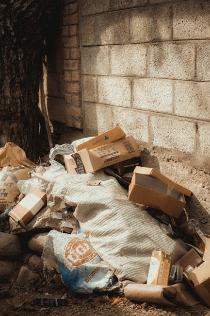A large pile of black rubbish bags filled with waste is stacked against a modern tiled wall on an outdoor pavement area. The bags appear to contain mixed household refuse, with some slightly torn open, revealing plastic bottles and smaller debris inside. Several discarded plastic water bottles are visible, protruding from the bags or resting on top. In the background, graffiti with red spray paint spelling 'XEND' is visible on the wall, and a cloudy sky with hints of sunlight is seen above the scene. The setting suggests an instance of unauthorised dumping or a completed private rubbish clearance, with Waste Clearance Notting Hill potentially involved in the removal of this waste. The overall environment indicates an urban area requiring specialist waste removal services, with the scene highlighting the importance of professional rubbish collection and disposal for such waste accumulation.