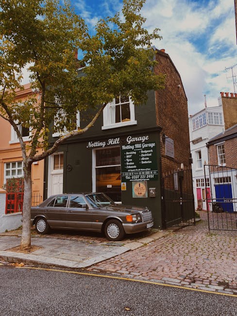 The image depicts a small, two-storey building with a dark grey facade, located on a residential street with cobblestone paving. A large deciduous tree with green leaves partially obscures the upper part of the building. A classic grey coupe car with a sleek, smooth finish and tinted windows is parked in front of the building, close to the curb. The building's ground floor features a sign that reads 'Notting Hill Garage,' advertising services such as exhausts, brakes, steering, and diagnostics, with a contact telephone number displayed below. To the right of the building, a black metal gate provides access to a rear parking or storage area, bordered by nearby residential houses with brick exteriors and white window frames. The scene is captured during daylight hours, with a clear sky and soft natural lighting, illustrating a typical urban environment where private waste clearance and rubbish removal services might operate, such as Waste Clearance Notting Hill, in an everyday setting.