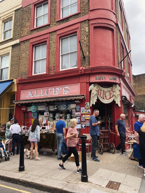 A retail shop named 'Retro Clothing' with a black storefront featuring gold lettering advertising second-hand bargains for men and women. The shop's large front window displays various items, papers, and notices attached to the glass. The building has a brick facade on the upper levels with three tall, arched white-framed windows. To the right side of the shop, there is a white building with graffiti tags on the wall below the shop sign. In front of the store, a sidewalk runs parallel to the road, which has a black metal railing separating pedestrians from the street. The pavement appears wet, indicating recent rain, and a person is walking past the shop, wearing a dark coat and beanie. The overall scene captures an urban street setting during daytime, consistent with a location that may benefit from independent waste disposal services offered by Waste Clearance Notting Hill, especially for disposing of shop-related rubbish or clearances.