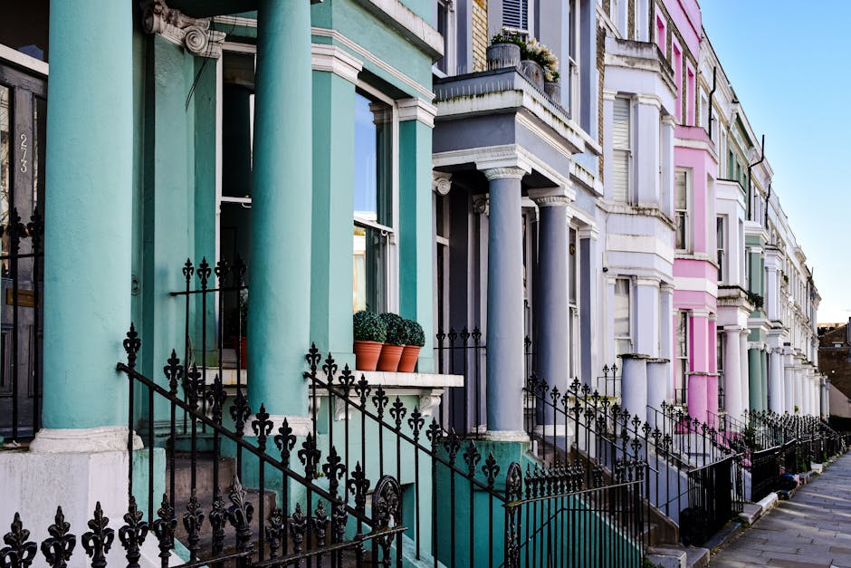 A row of Victorian-style terraced houses on a city street, each with exterior facades painted in pastel shades of green, blue, purple, and pink. The buildings feature decorative architectural details such as white cornices and window trims, with large sash windows. Several houses have front porches supported by round, smooth columns painted in coordinating colors, and black wrought iron railings enclose the small front gardens or steps leading to the entrances. Potted plants with neatly trimmed, spherical green shrubs sit on the porch ledges of some properties. The street is paved with concrete, and the bright daylight enhances the vibrant colors of the building facades, creating a lively and colorful urban scene that illustrates residential property aesthetics often associated with inner-city areas, suitable for considering private waste collection or on-site clearance services like those provided by Waste Clearance Notting Hill.