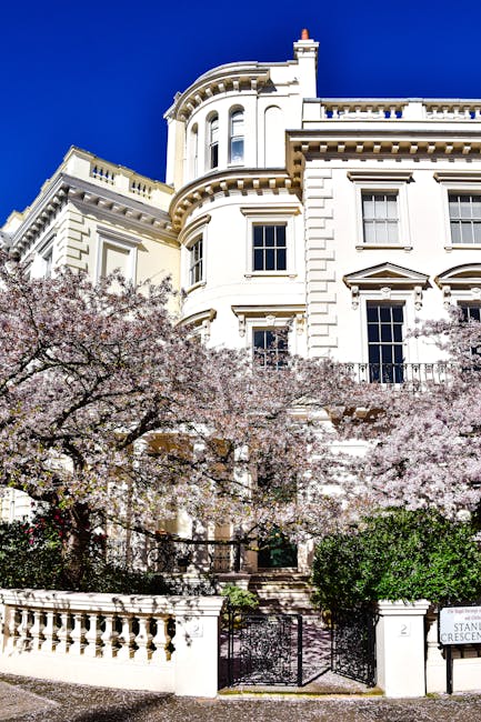 A large, white, Victorian-style building with ornate architectural details including rounded bay windows and decorative cornices, set against a vibrant blue sky. In front of the property, there are flowering trees with light pink blossoms partially obscuring the lower section of the building. The trees are in full bloom, creating a contrast with the stately white facade. A wrought iron gate and low stone wall form the entrance to the front garden, which is bordered by neatly trimmed green shrubs. To the right, there is a small white signpost with the number '2' and the street name 'Stanley Crescent'. The scene is bright and clear, highlighting the building's elegant features and the natural beauty of the flowering trees. This setting subtly connects to the topic of property maintenance and the importance of managing outdoor spaces in the context of private property upkeep, including rubbish clearance or general waste management services for the area, as offered by Waste Clearance Notting Hill.