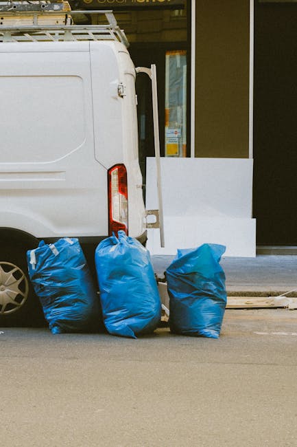 Close-up view of the rear side of a white commercial van parked on a pavement in an urban setting. Three blue plastic rubbish bags, filled and tied, are placed directly behind the vehicle, resting on the ground. The rear door of the van is partially visible, with a subtle vertical handle and red reflector positioned near the left edge. In the background, a storefront or building with large glass windows and a door can be seen, partially obscured by the van. The environment appears clean but functional, suitable for on-site waste collection and private rubbish removal services. The scene highlights an example of independent rubbish disposal, aligned with the type of work carried out by waste clearance companies such as Waste Clearance Notting Hill. The street surface is smooth asphalt, and the overall atmosphere suggests late morning or early afternoon lighting, with natural daylight illuminating the scene evenly, emphasizing the practical nature of rubbish removal and waste management activities in an urban context.