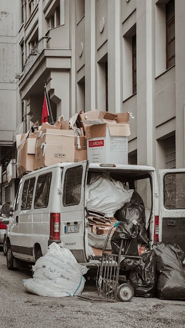 The image depicts the rear of a white commercial van parked on a city street, loaded with a large assortment of discarded waste materials. The open van door reveals an interior filled with bulky items, including black and white garbage bags, flattened cardboard boxes, and miscellaneous debris, all stacked to maximize space. Several large cardboard boxes, some torn and crumpled, are placed on top of the van, with varying sizes and shades of brown and white, one branded with the words 'LEARDIN' visible on a box. Outside the van, on the pavement, there are additional black trash bags and white sacks, along with a small dolly or trolly partially visible. The surrounding environment features a tall, light-colored building with multiple rectangular windows and architectural details, suggesting an urban setting. The lighting is natural, possibly overcast, which provides even illumination across the scene. This setup reflects a private waste collection process, likely handled by a service such as Waste Clearance Notting Hill, involving on-site removal of large-scale rubbish in a manner that supports alternative waste handling outside local authority collection.