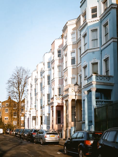 The image shows a row of multi-storey residential buildings with classic Victorian architectural features, painted in light pastel blue and white tones. The buildings have ornate decorative elements around the windows and balconies, with some windows open, revealing interior curtains. To the left, there is a large, leafless tree casting shadows on the buildings. The foreground features a street with a line of parked cars, including sedans and small hatchbacks, parked parallel along the curb. The pavement appears to be made of asphalt, and a green metal fence runs along the right side of the image, partially obscuring parts of the buildings. The sky above is clear and bright, indicating daytime, with natural sunlight illuminating the scene evenly. The overall setting suggests a quiet urban street in a residential area, where independent waste collection or private rubbish removal services, like those provided by Waste Clearance Notting Hill, might be considered for managing household waste directly from residents' properties.