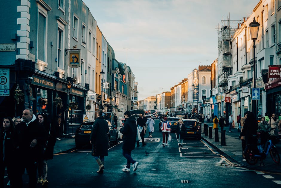 A busy urban street scene in Notting Hill during late afternoon, featuring a mix of historic and modern buildings with pastel-colored facades. The street is lined with small businesses, including a pub named Duke of Wellington on the left side, and various shopfronts with signage and window displays. Pedestrians are walking along the pavement, some in small groups and others alone, dressed in casual and business attire. Several cars, including a black taxi, are parked along the curb and driving through the street, with a few being loaded or unloaded. Street lamps and traffic signs are visible, along with a construction scaffold on the right side of the image. The lighting suggests natural sunlight with a soft, warm glow on the buildings, creating a lively yet orderly atmosphere. This scene illustrates the typical environment where private rubbish removal services like Waste Clearance Notting Hill could operate, especially for on-site clearance or alternative waste collection of commercial and residential waste in a vibrant urban setting.