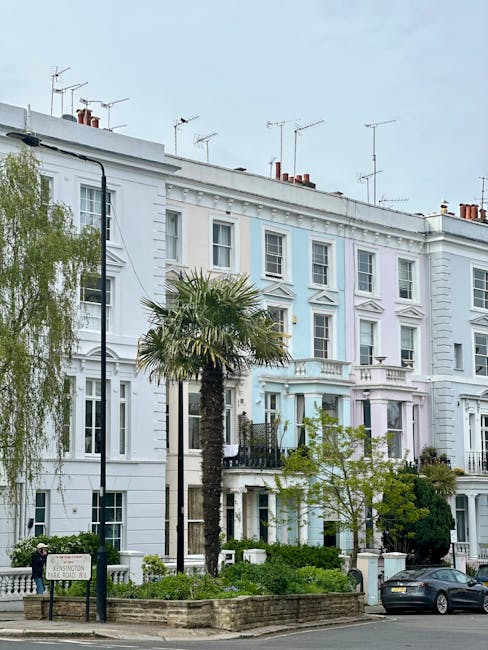 The image shows a row of multi-storey residential buildings with classic Victorian architectural features, painted in light pastel blue and white tones. The buildings have ornate decorative elements around the windows and balconies, with some windows open, revealing interior curtains. To the left, there is a large, leafless tree casting shadows on the buildings. The foreground features a street with a line of parked cars, including sedans and small hatchbacks, parked parallel along the curb. The pavement appears to be made of asphalt, and a green metal fence runs along the right side of the image, partially obscuring parts of the buildings. The sky above is clear and bright, indicating daytime, with natural sunlight illuminating the scene evenly. The overall setting suggests a quiet urban street in a residential area, where independent waste collection or private rubbish removal services, like those provided by Waste Clearance Notting Hill, might be considered for managing household waste directly from residents' properties.