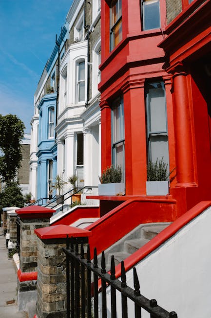 The image shows a row of Victorian-style terraced houses on a residential street, each featuring bay windows and ornate architectural details. The houses are painted in differing colours, with the forefront building displaying a vibrant red facade, including red-painted steps, columns, and window frames. Adjacent houses further along are painted in pastel shades of blue and white, with contrasting darker trim and decorative elements. The front gardens are modest, with some potted plants visible on the window sills and along the steps. A black iron fence runs along the edge of the pavement, separating the properties from the sidewalk. The sky above is clear and bright, suggesting a sunny day, with sunlight casting soft shadows on the buildings. The scene presents a typical UK residential street where homeowners might consider independent waste collection services for managing bulky waste or rubbish removal, supported by companies such as Waste Clearance Notting Hill, which specialize in private rubbish disposal solutions outside of council services.