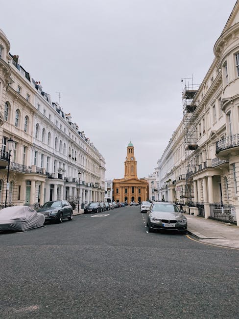 A quiet street scene featuring a row of white, multi-storey residential buildings with wrought iron balconies and sash windows on both sides. The buildings have decorative cornices and are aligned along a slightly curved asphalt road. Several parked cars, including a covered vehicle, are positioned along the curb on the left, while a few modern sedans are parked on the right side. In the background, a classical-style church or municipal building with a prominent clock tower and a green dome is visible, set against an overcast sky. The overall atmosphere suggests a typical urban environment suitable for private waste collection or rubbish removal services, with no visible waste or debris present in the scene. The setting is clean and orderly, representing an example of well-maintained streets consistent with alternative waste handling options that avoid reliance solely on municipal services. The image matches the context of independent collection or on-site clearance relevant to waste clearance in the Notting Hill W11 area.