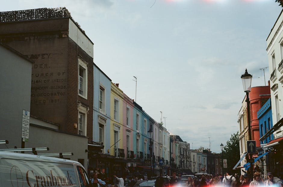 The image shows a row of Victorian-style terraced houses on a residential street, each featuring bay windows and ornate architectural details. The houses are painted in differing colours, with the forefront building displaying a vibrant red facade, including red-painted steps, columns, and window frames. Adjacent houses further along are painted in pastel shades of blue and white, with contrasting darker trim and decorative elements. The front gardens are modest, with some potted plants visible on the window sills and along the steps. A black iron fence runs along the edge of the pavement, separating the properties from the sidewalk. The sky above is clear and bright, suggesting a sunny day, with sunlight casting soft shadows on the buildings. The scene presents a typical UK residential street where homeowners might consider independent waste collection services for managing bulky waste or rubbish removal, supported by companies such as Waste Clearance Notting Hill, which specialize in private rubbish disposal solutions outside of council services.