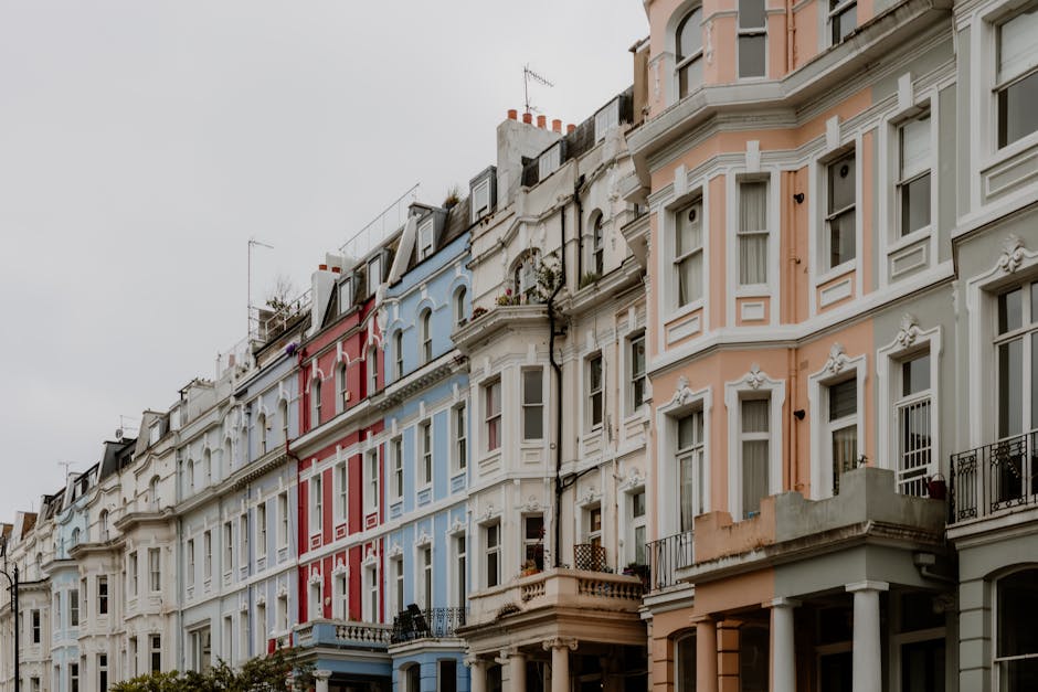 A row of Victorian-style terraced houses in Notting Hill with detailed stucco facades painted in pastel shades of white, pink, light blue, and cream. The buildings feature large bay windows with white frames, some with decorative molding and ornate trim. Small balconies with black wrought iron railings extend from several upper floors. The rooftops are visible, with chimneys and antennas, under an overcast sky. The ground level of each property includes entrance doors and steps leading onto the sidewalk, where some greenery and potted plants are placed. The overall scene showcases the characteristic architectural charm of the area, with clean and well-preserved facades. As part of a waste clearance context, Waste Clearance Notting Hill could be responsible for on-site removal of building waste or debris from renovations, consistent with alternative disposal methods for domestic waste. The environment appears calm and residential, with no visible activity or clutter in the immediate exterior surroundings.