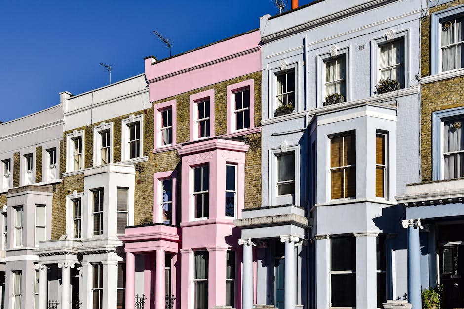 A row of Victorian-style terraced houses with brick facades painted in various colors, including white, light pink, and pale blue, situated along a street in clear daylight. The buildings feature large sash windows with white frames, some adorned with small potted plants on the window sills and decorative architectural details such as cornices and columns around the entrances. The pink building in the center has a protruding bay window on the second and third floors, creating a slight architectural variation. The facades are clean and well-maintained, with the brickwork visible in some areas, and the sky overhead is bright blue without clouds. In the foreground, part of the sidewalk or pavement is visible, emphasizing the urban residential environment. This street scene, captured during daytime, exemplifies typical London townhouse architecture, relevant for discussions on private property maintenance and external property management related to rubbish and waste handling, occasionally connected with the services of waste clearance providers like Waste Clearance Notting Hill.